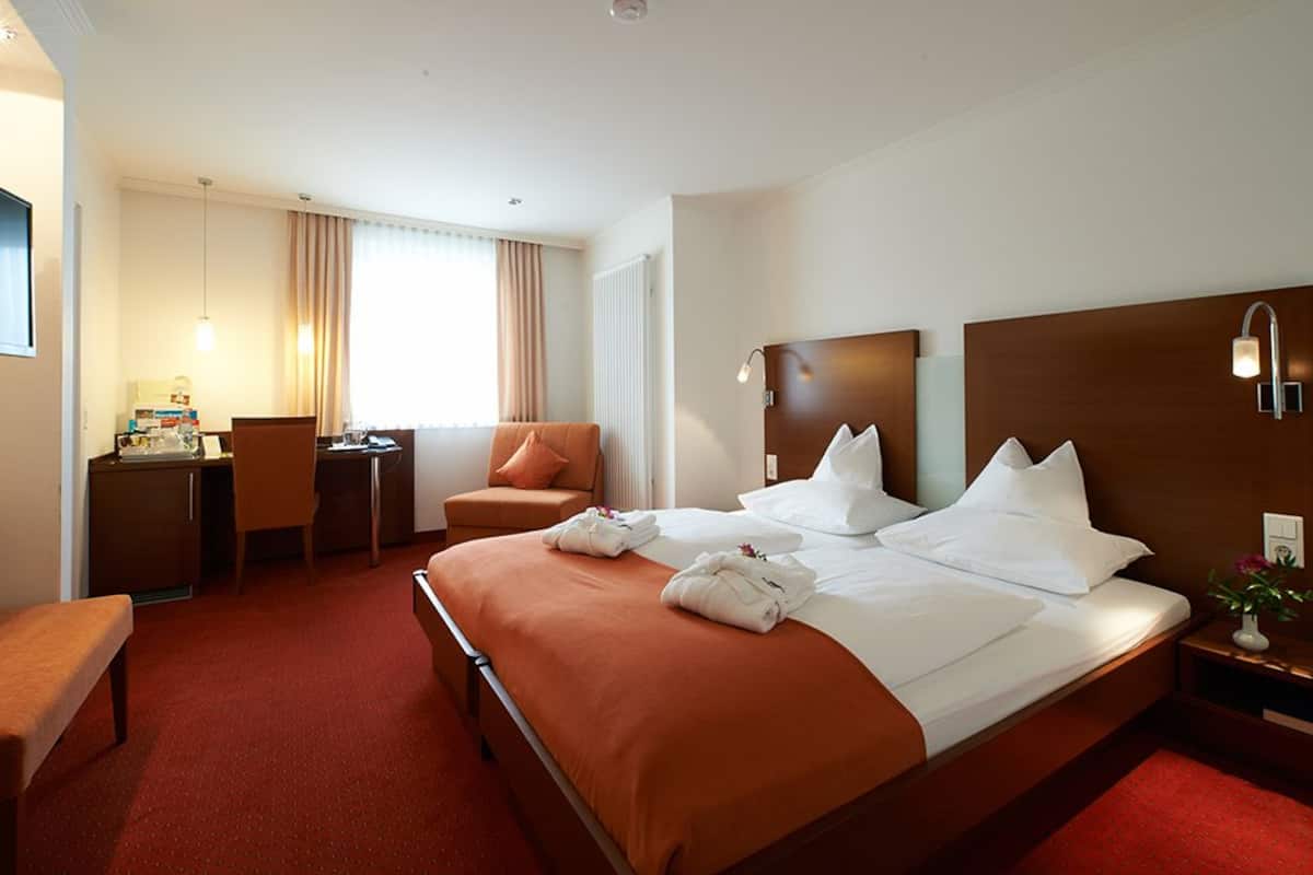 A hotel room with a large bed, a desk, a chair, and a small table with a lamp.