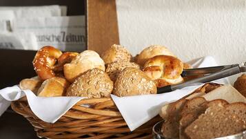 A basket of various breads and rolls, including some with sesame seeds, on a table with a newspaper in the background.