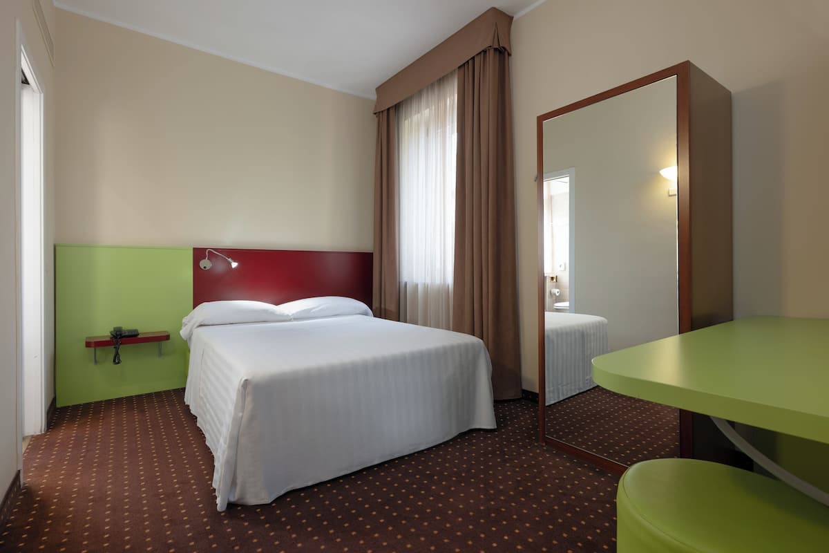 Quarto de hotel com uma cama de solteiro, uma mesa de cabeceira verde, um espelho e uma cadeira verde.