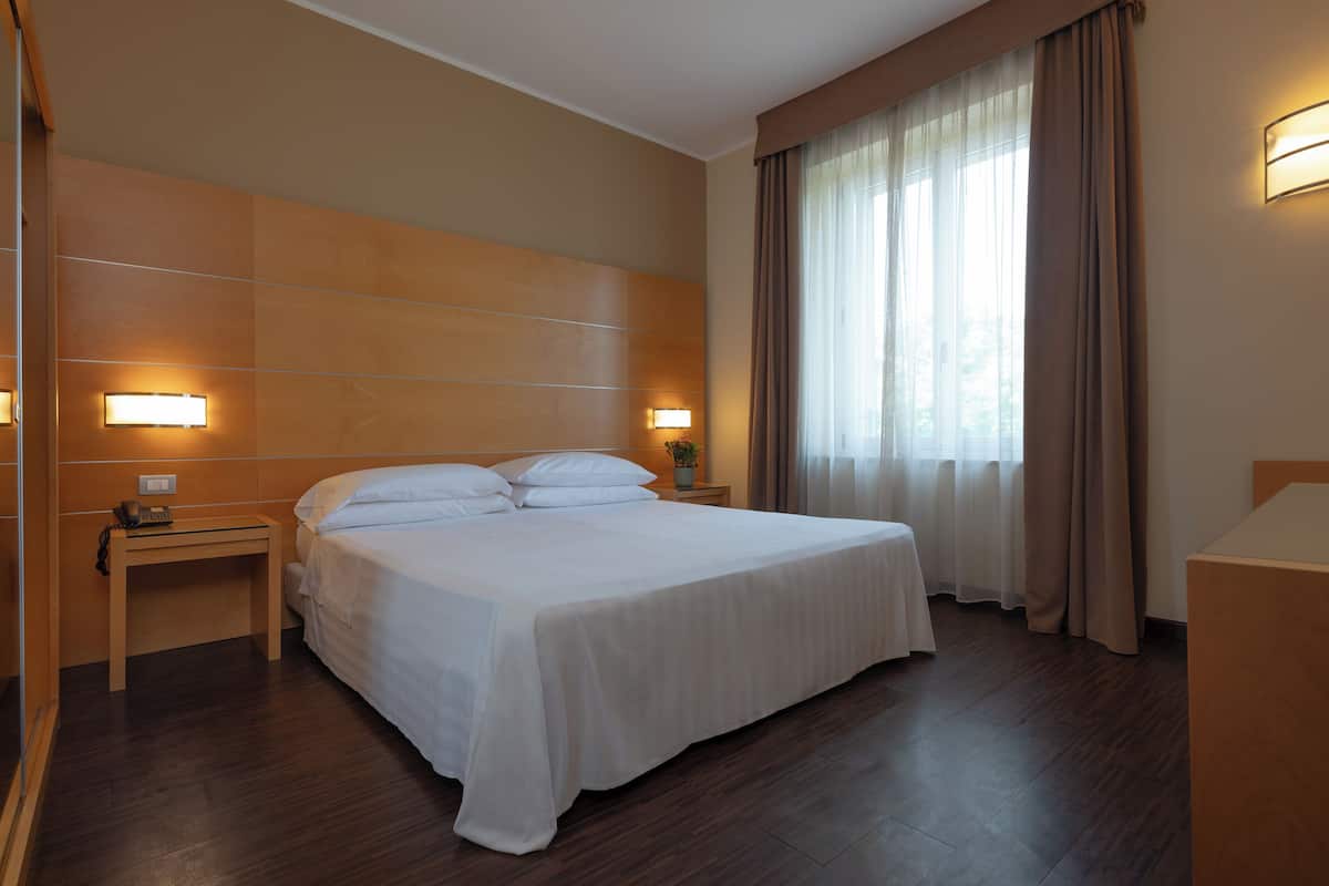 Quarto de hotel com uma cama grande, luminárias de cabeceira, mesa de cabeceira e janela com cortinas.