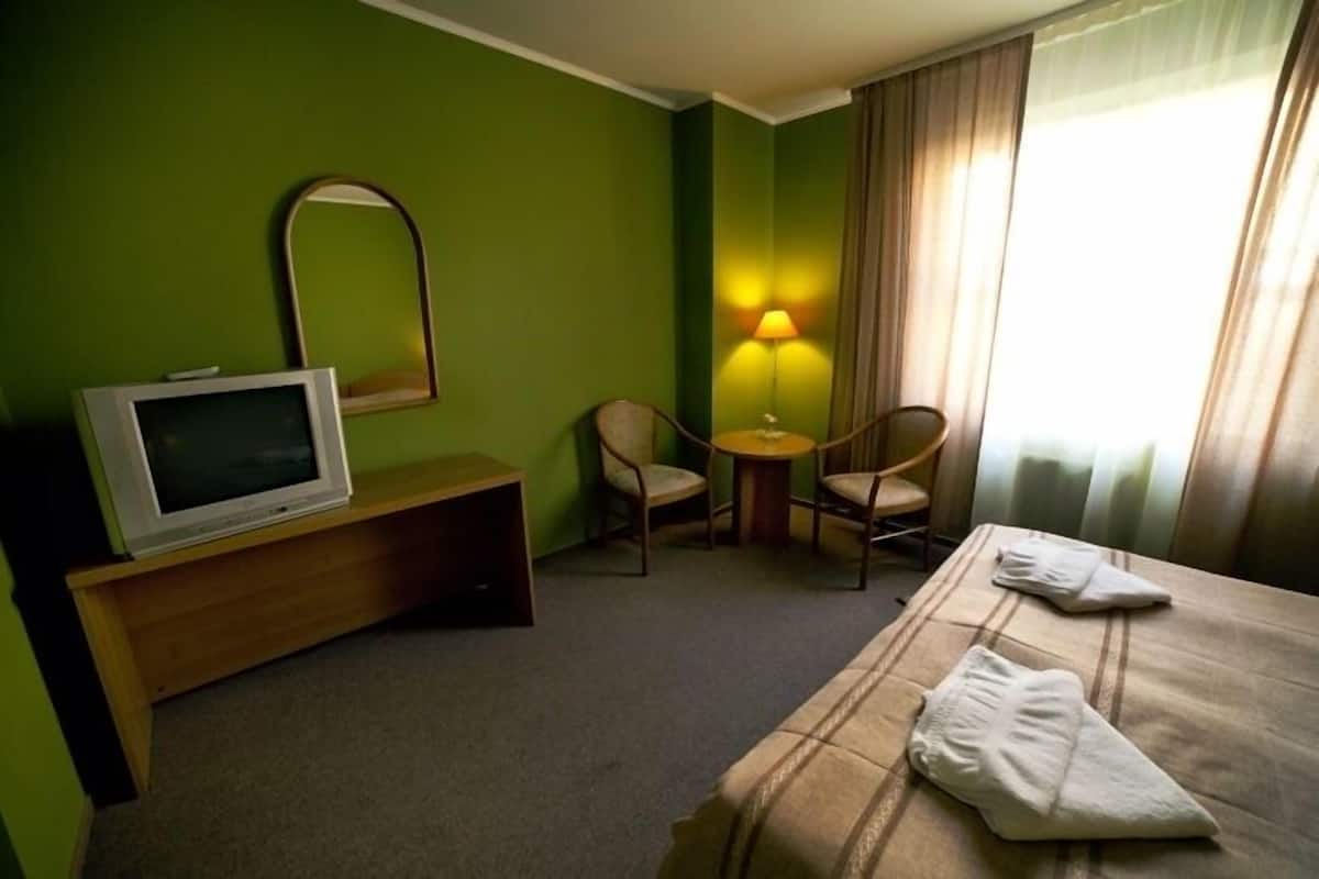 Une chambre d’hôtel avec un lit, un téléviseur posé sur un meuble en bois, un bureau, deux chaises, une lampe et une fenêtre avec des rideaux.