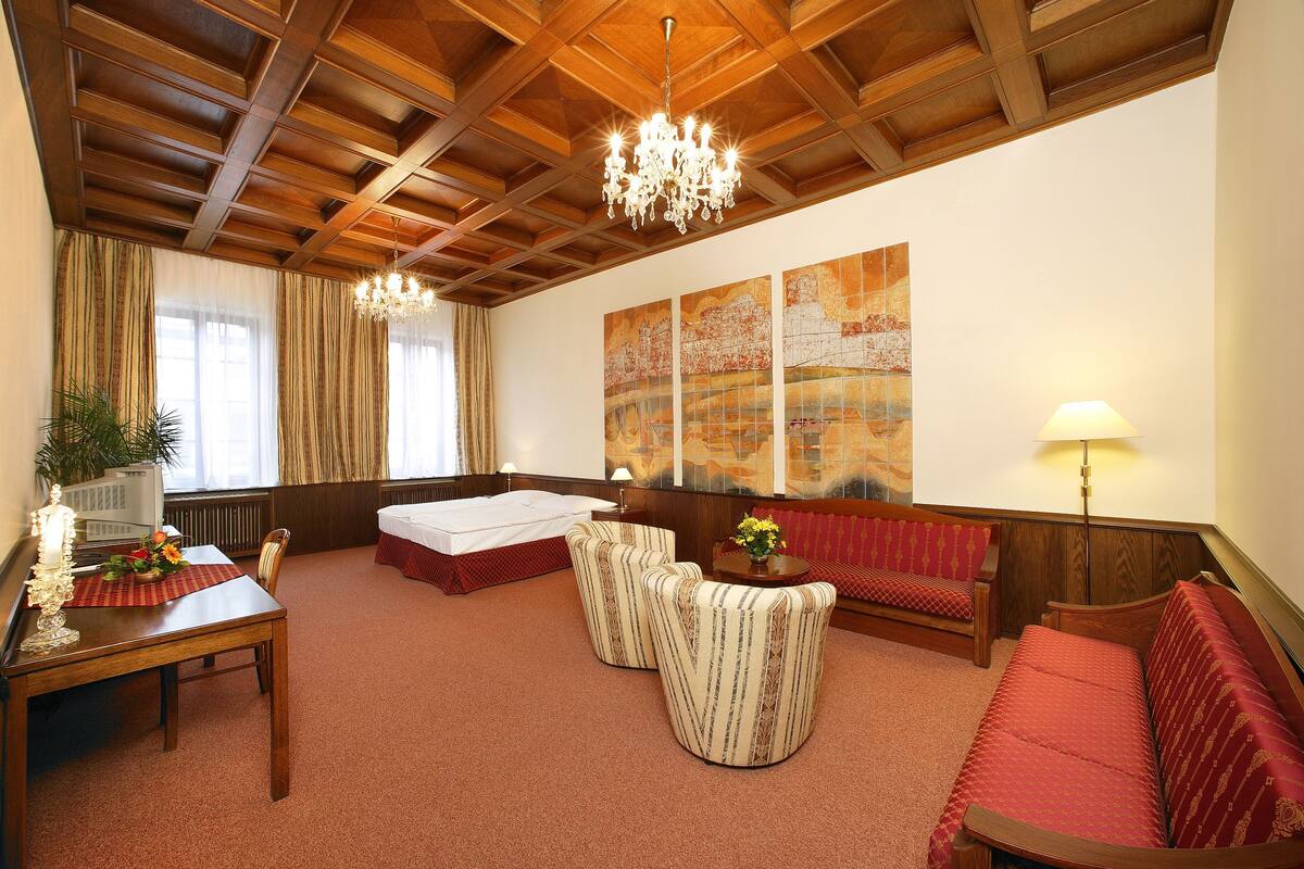 A hotel room with a wooden ceiling, a chandelier, a bed, a red sofa, a small table with a lamp, and a large abstract painting on the wall.