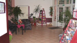 Una hall con pavimento piastrellato, un divano in pelle nera, una poltrona rosa, una libreria a scala in legno con piante e un espositore per riviste.