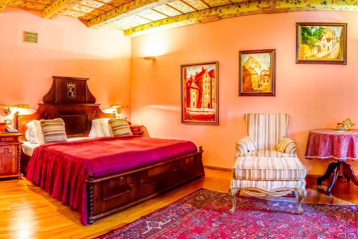 A bedroom with a wooden bed, a piano, a chair, a table, and two paintings on the wall.