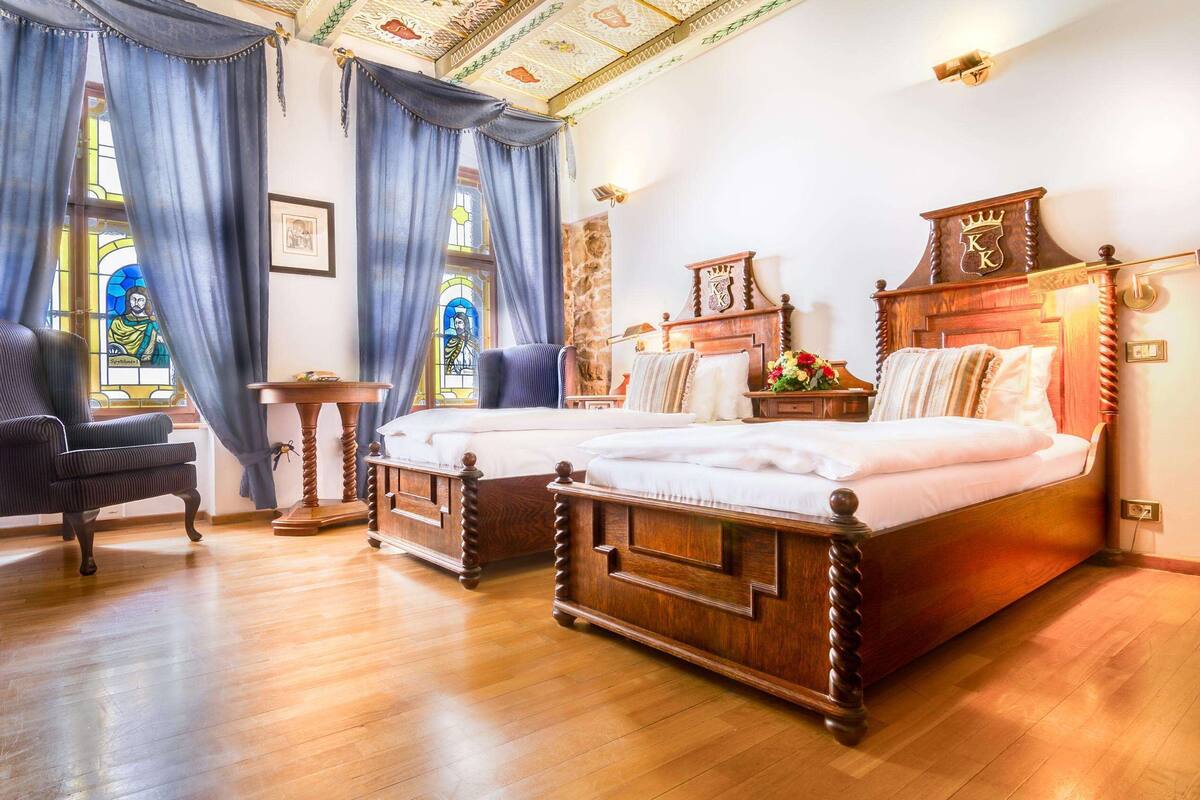 A traditional bedroom with two wooden beds, a chair, a small table, and a large window with stained glass.