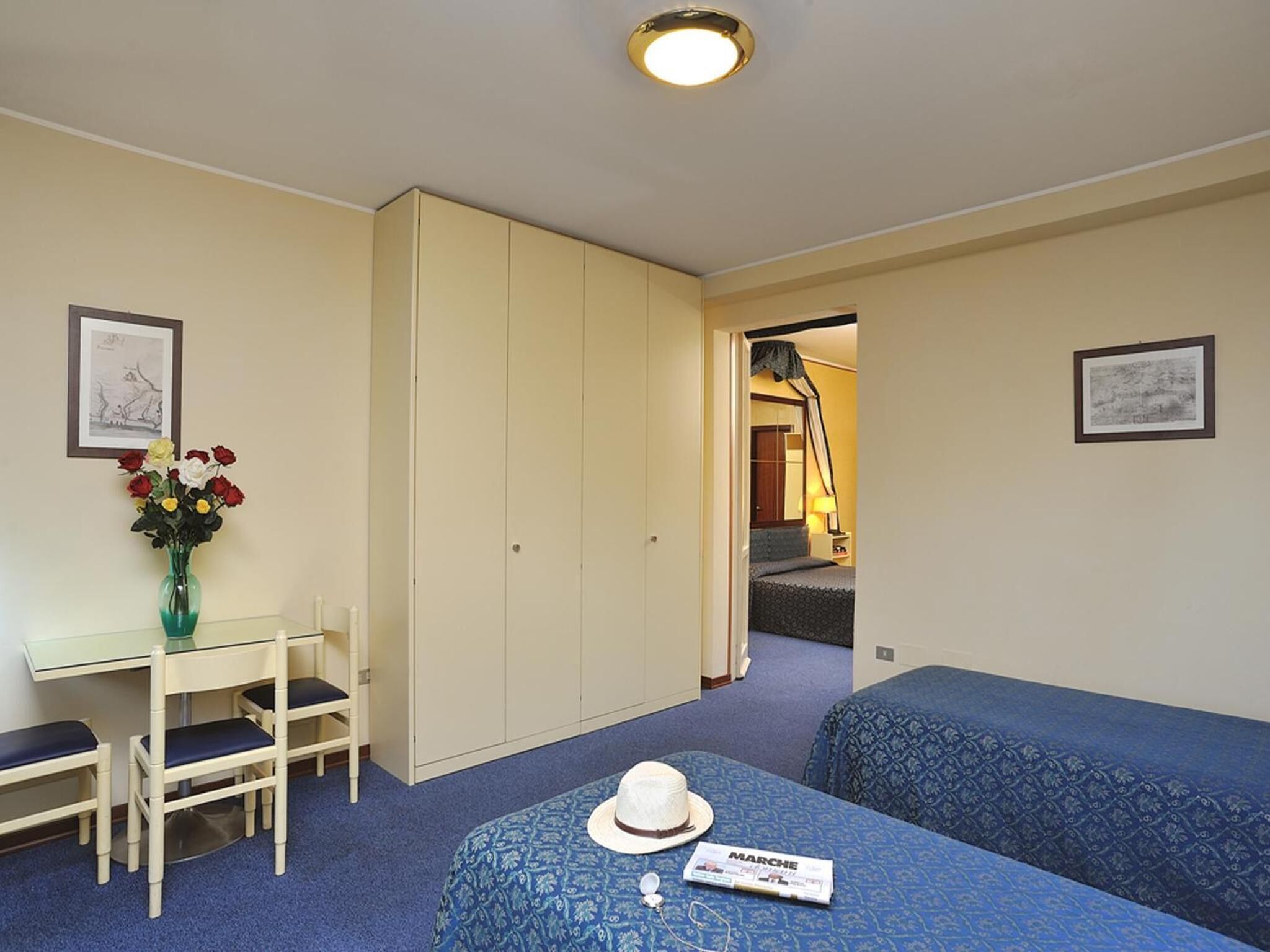 Een kamer met een bed, een bureau met stoelen, een vaas met bloemen en een kledingkast.