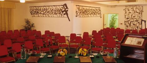 Un salón de conferencias con filas de sillas rojas, una larga mesa con centros de mesa florales y arte abstracto en las paredes.