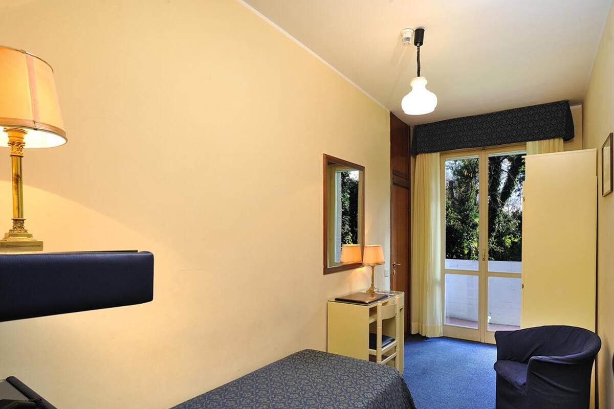 Ein Hotelzimmer mit Bett, Schreibtisch, Stuhl, Lampe und Fenster mit Blick auf Grünflächen.