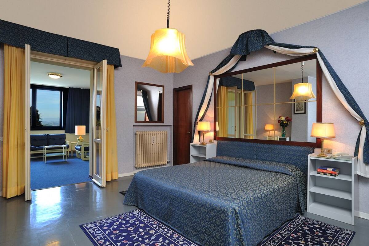 Ein Hotelzimmer mit einem großen Bett, einer Sitzecke mit Sofa und Sesseln, einem Esstisch und Blick ins Freie.