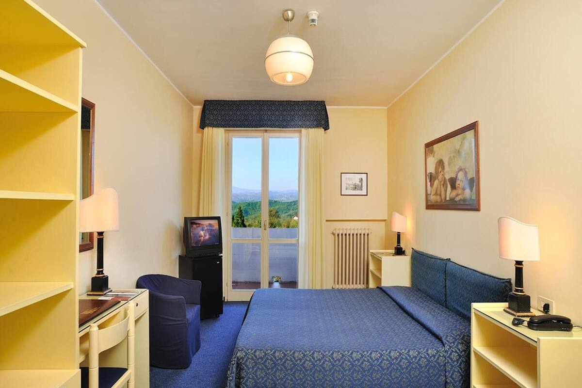 Ein Hotelzimmer mit Bett, Schreibtisch, Stuhl, Fernseher, einem Fenster mit Aussicht und einem Bild an der Wand.