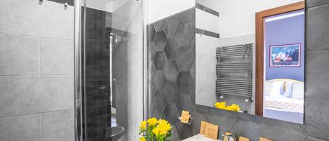 A modern bathroom with a glass shower enclosure, a large sink with toiletries, and a mirror reflecting a bed with a blue wall.
