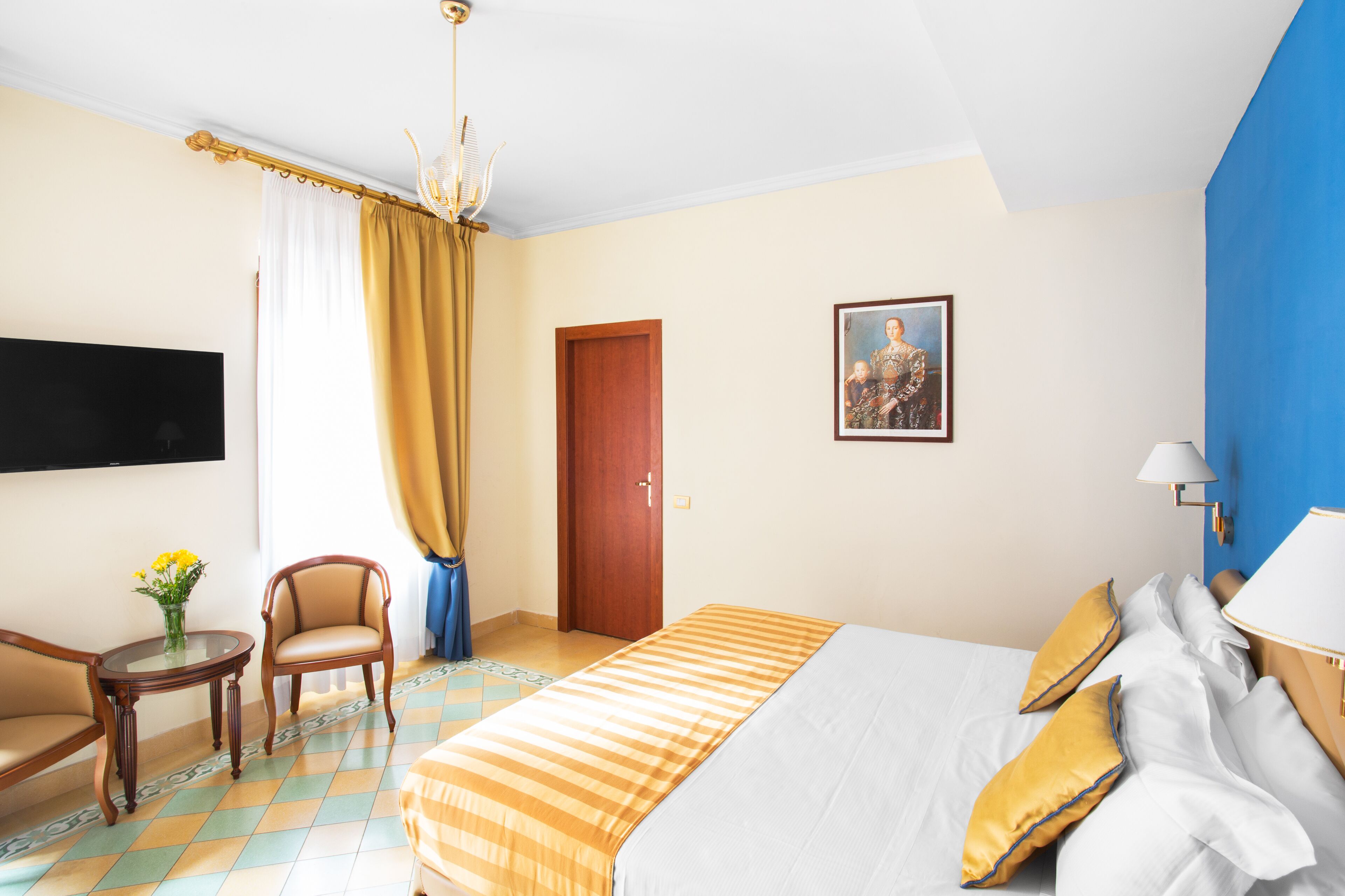 A hotel room with a bed, a chair, a table with a vase, a painting, and a television on the wall.