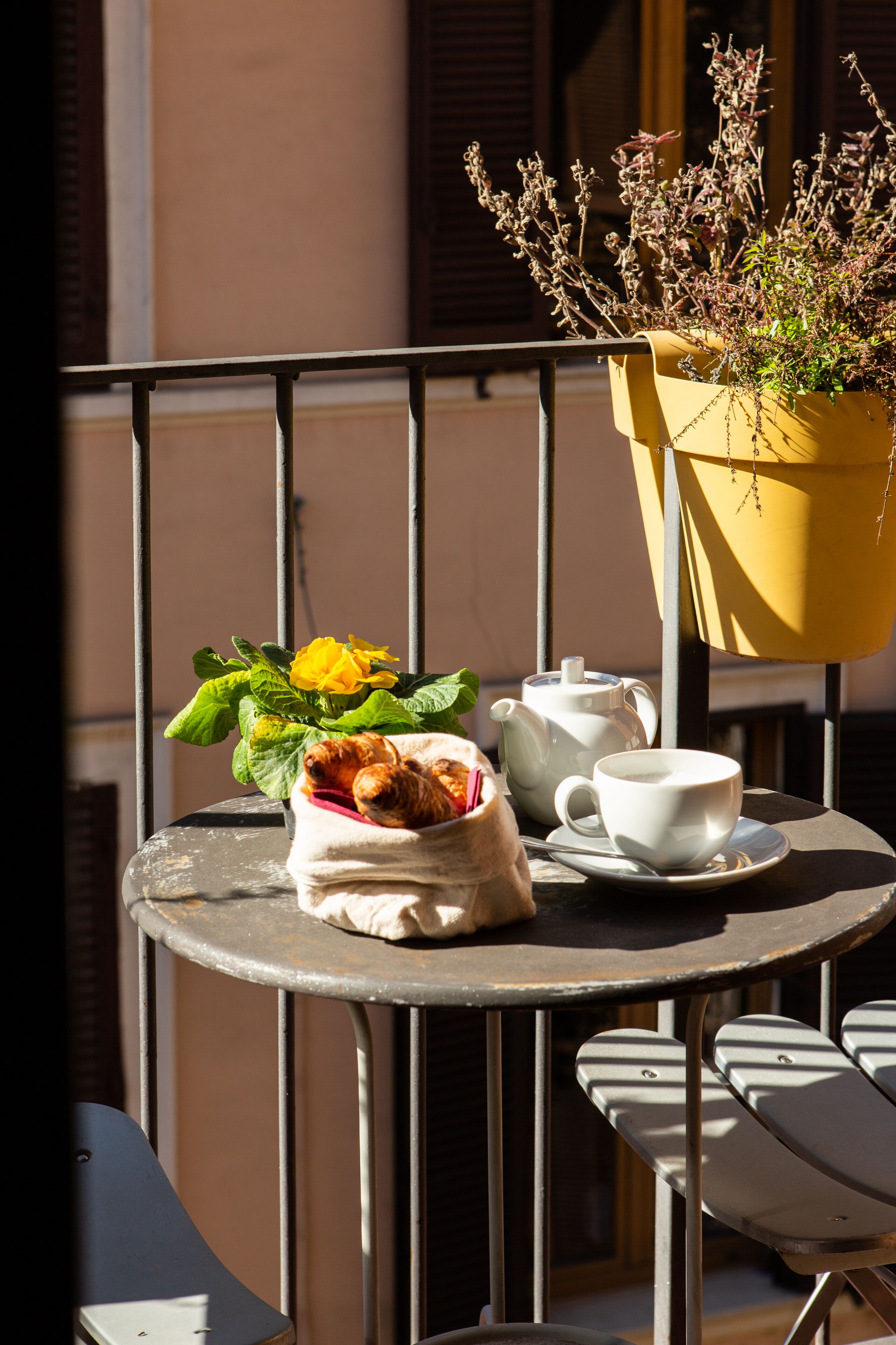 A round metal table with a white teapot and cup, a small bag of pastries, and a yellow planter with dried plants.