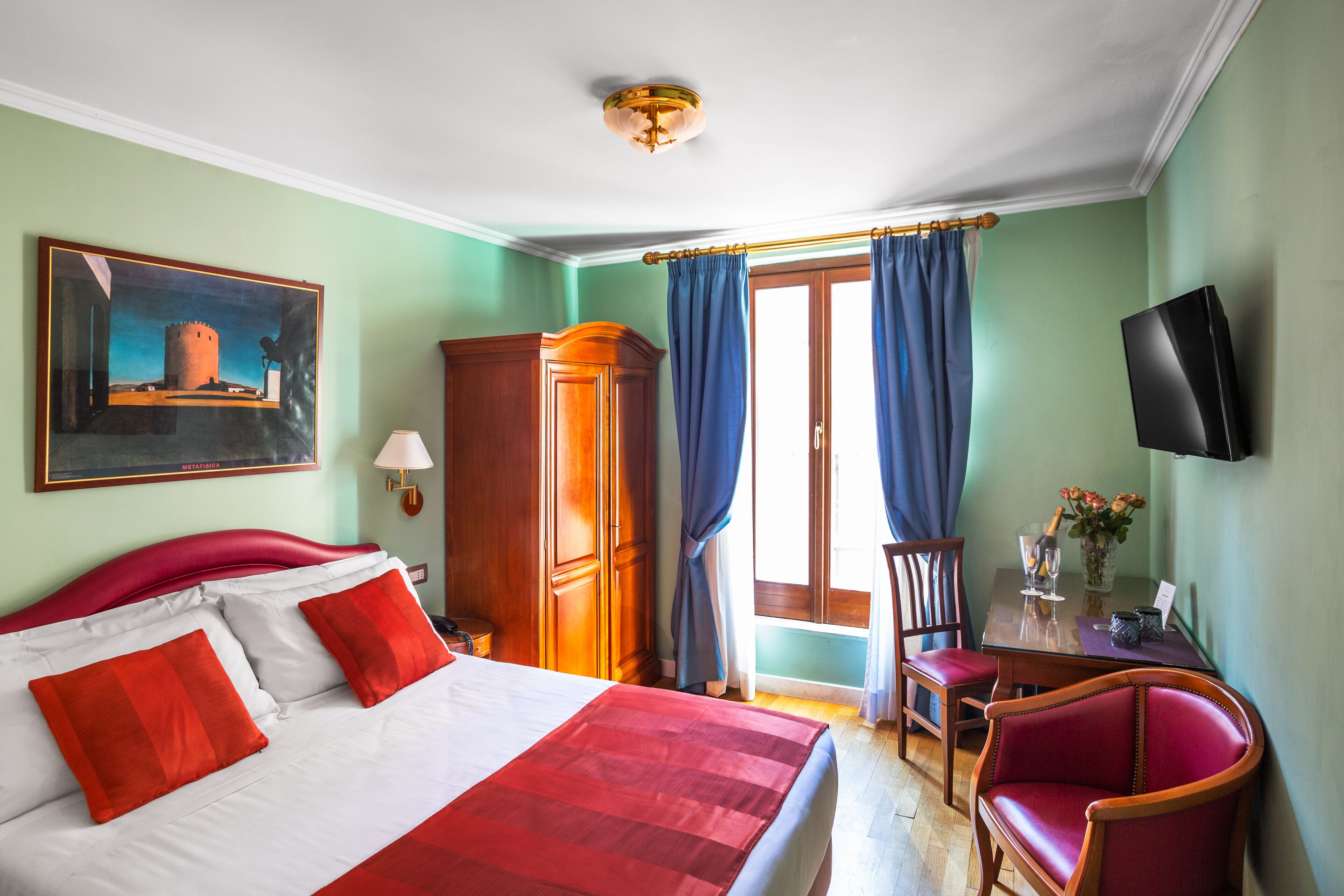 A hotel room with a bed, a desk, a chair, a television, and a painting on the wall.