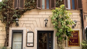 A hotel facade with a traditional design, featuring a ground-floor entrance with a black door, a signboard above the door, and a window with shutters. There are potted plants in front of the entrance and a signboard to the right. - Hotel Piazza Di Spagna (Rome)