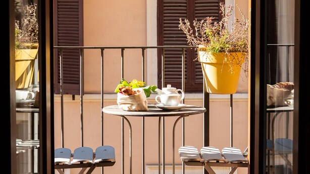 A balcony with a table and chairs, potted plants, and a view of a building with shutters. - Hotel Piazza Di Spagna (Rome)
