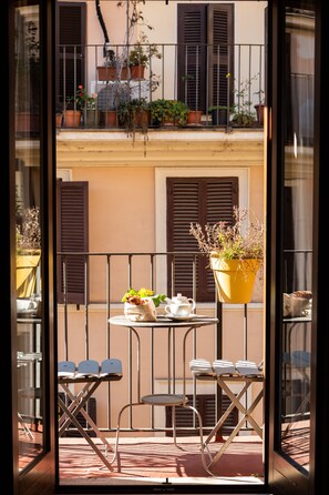 A balcony with a table and chairs, potted plants, and a view of a building with shutters. - Hotel Piazza Di Spagna (Rome)