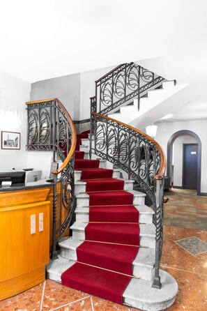 Una imponente escalera con barandales de metal ornamentados y peldaños alfombrados en rojo, que conduce a una zona de recepción con un mostrador de madera.