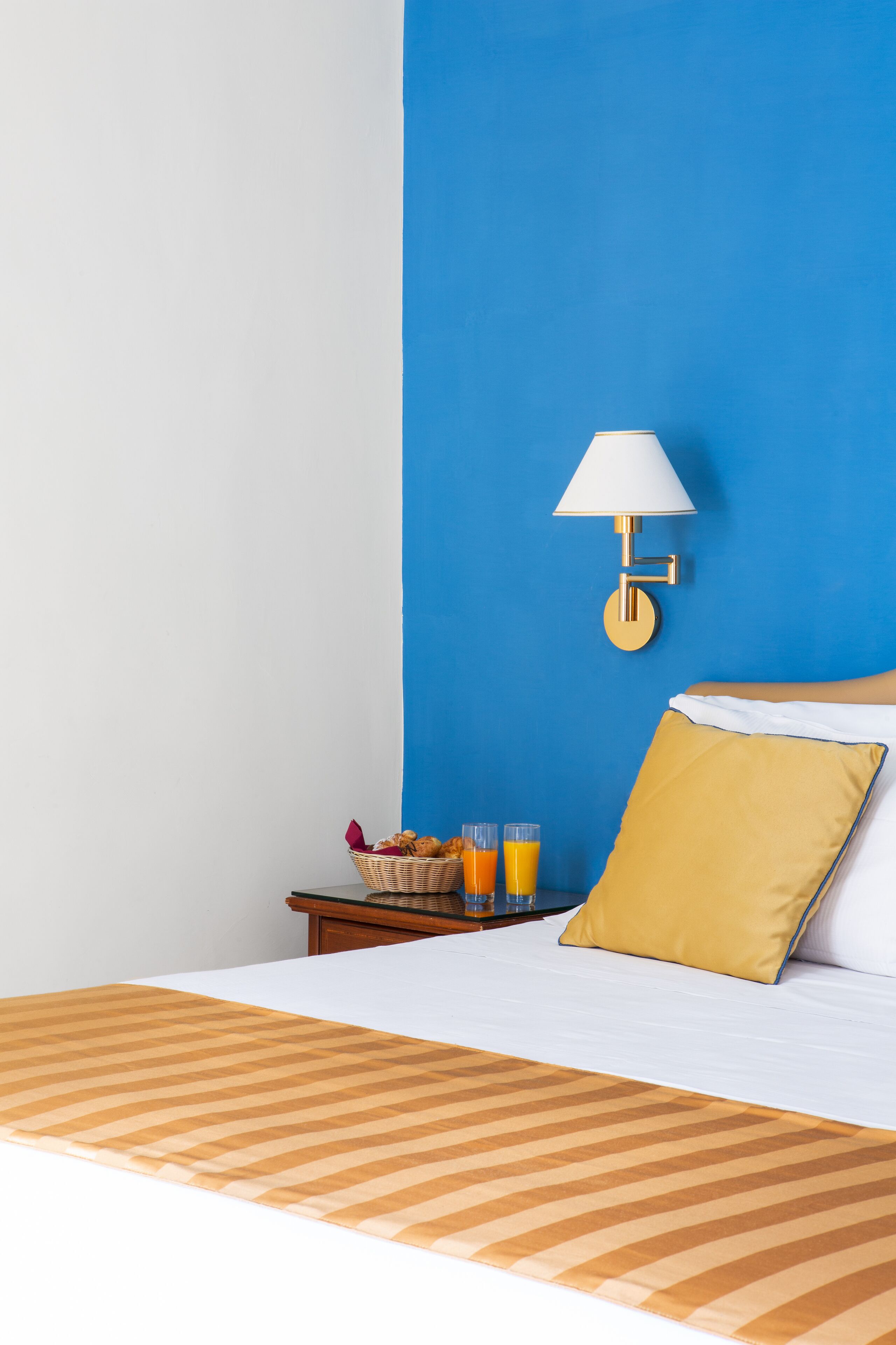 A hotel room with a blue accent wall, a bedside table with a basket of fruits and glasses of orange juice, and a bed with a yellow pillow.
