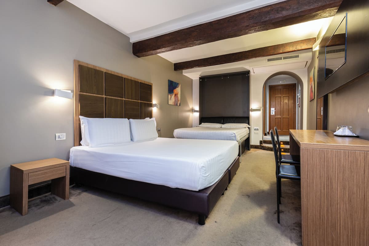 A hotel room with a large bed, a desk, and a chair. There is a wooden headboard and a small bedside table.