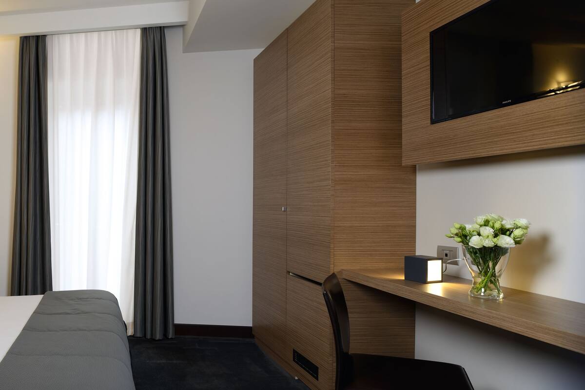 A modern hotel room with a large bed, a bedside table with a phone, a flat-screen TV mounted on a wooden panel, and a vase with flowers on a nightstand.