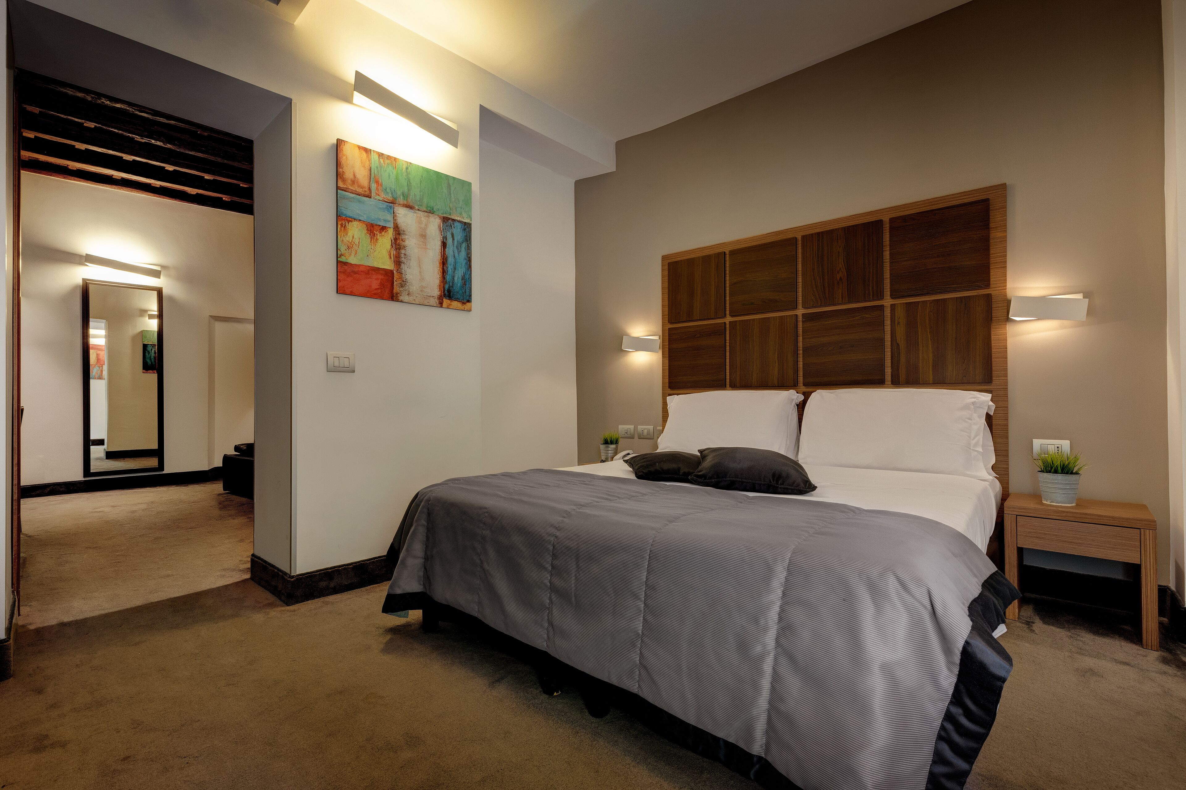 A bedroom with a large bed, a wooden headboard, a bedside table with a plant, and a colorful abstract painting on the wall.