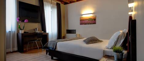 A bedroom with a bed, a desk, a chair, a TV, a painting, and a bedside table with a plant.