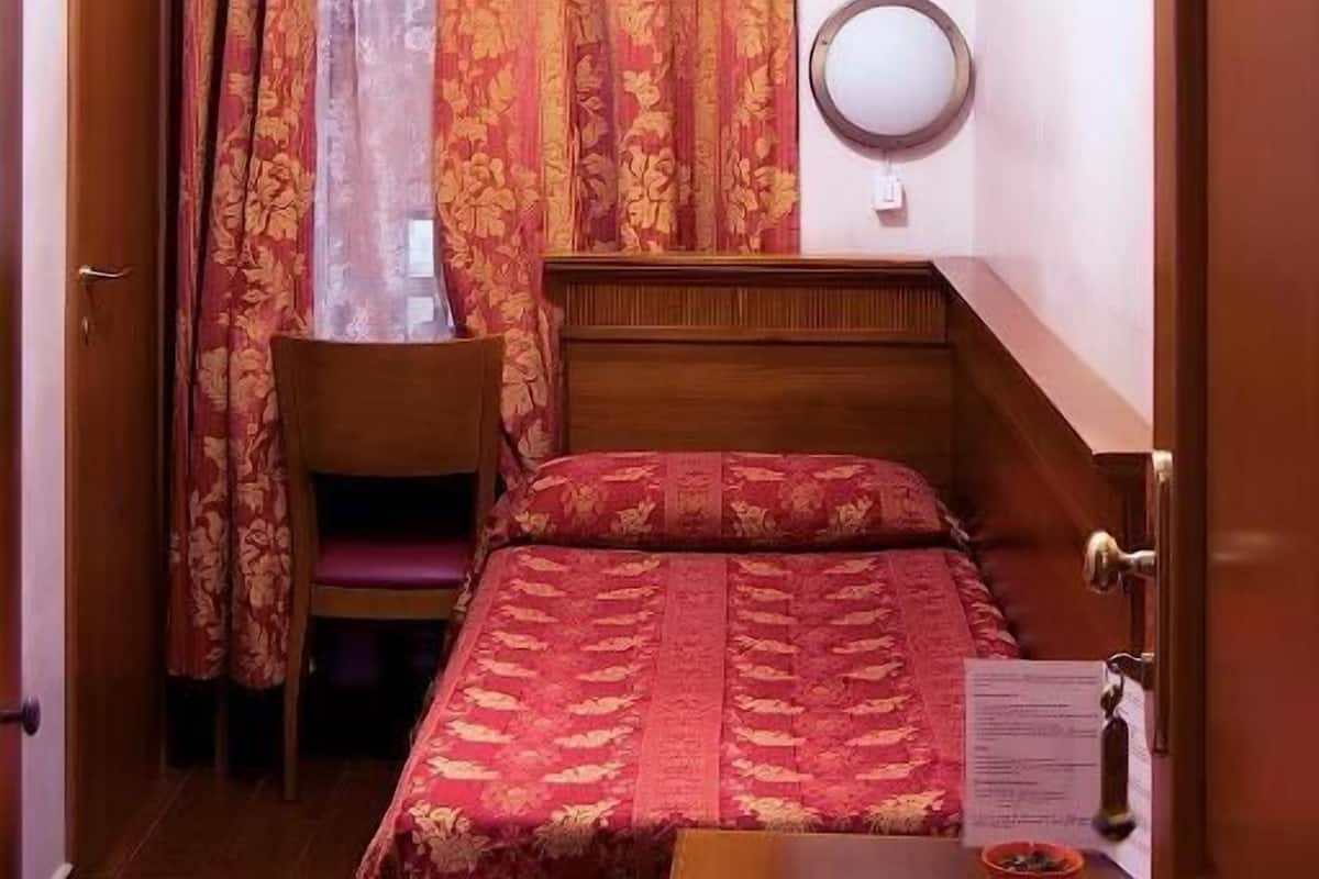 A hotel room with a bed, a wooden nightstand, a chair, a desk, a mirror, and a window with curtains.