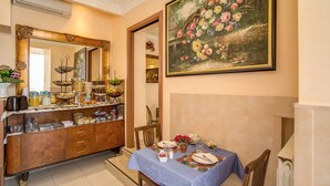 Uma área de refeições com uma mesa posta para dois, um buffet com diversos alimentos e um grande quadro floral na parede.