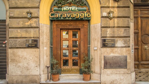 Entrada de um hotel com porta de madeira, duas plantas em vasos e uma placa acima da porta com a inscrição "HOTEL Caravaggio PICASSO".