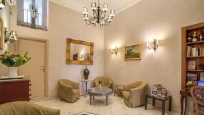 Uma sala de estar bem iluminada com sofás bege, uma mesa de centro redonda, um candelabro, uma estante de livros e um quadro na parede.