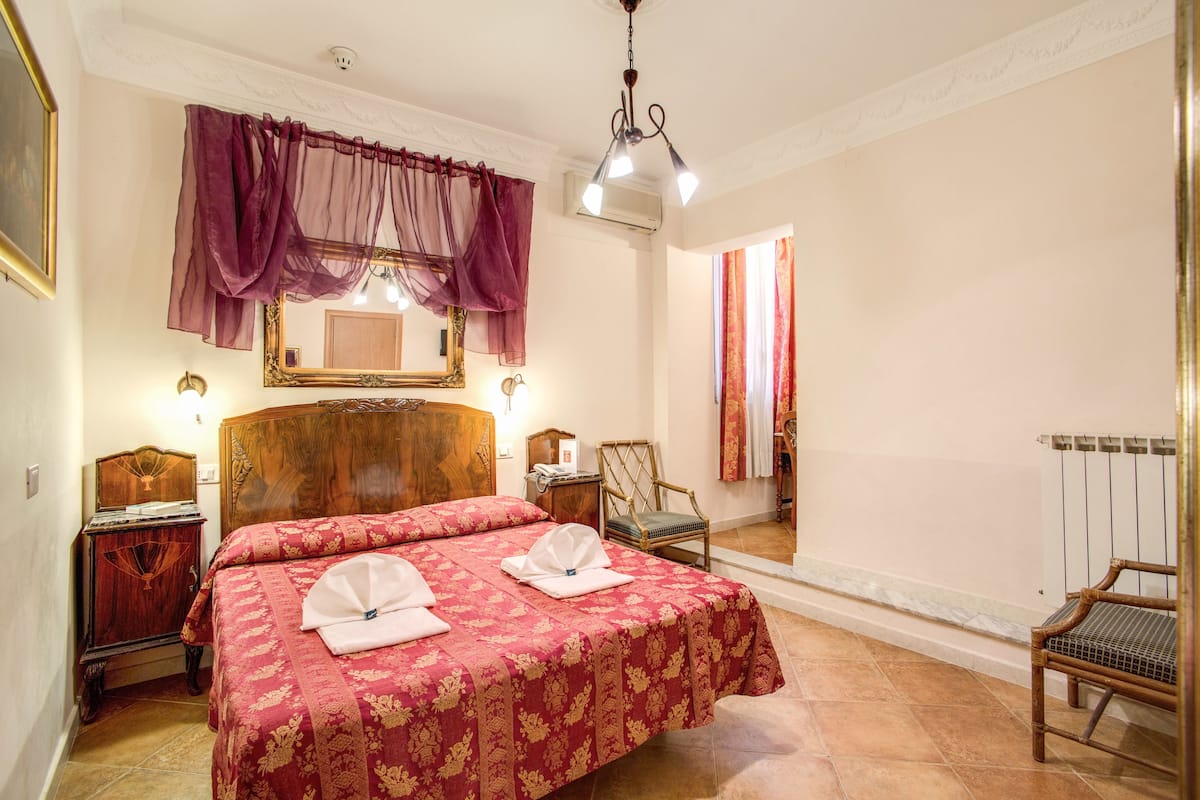 A hotel room with a large bed, a wooden headboard, a chair, a nightstand, a mirror, and a window with curtains.