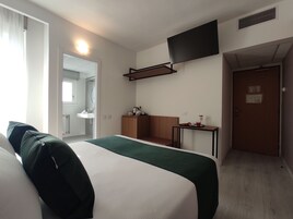 Camera d'albergo con un letto, un tavolino con una tazza e un libro, una TV a parete e una porta che dà sul bagno.