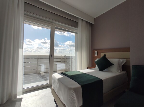 A hotel room with a bed, a nightstand, a phone, and a view of the sky and clouds through a sliding glass door.