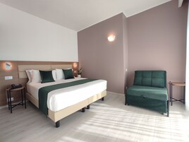 Et hotelværelse med en seng, en grøn lænestol, et lille bord og en vægmonteret lampe.