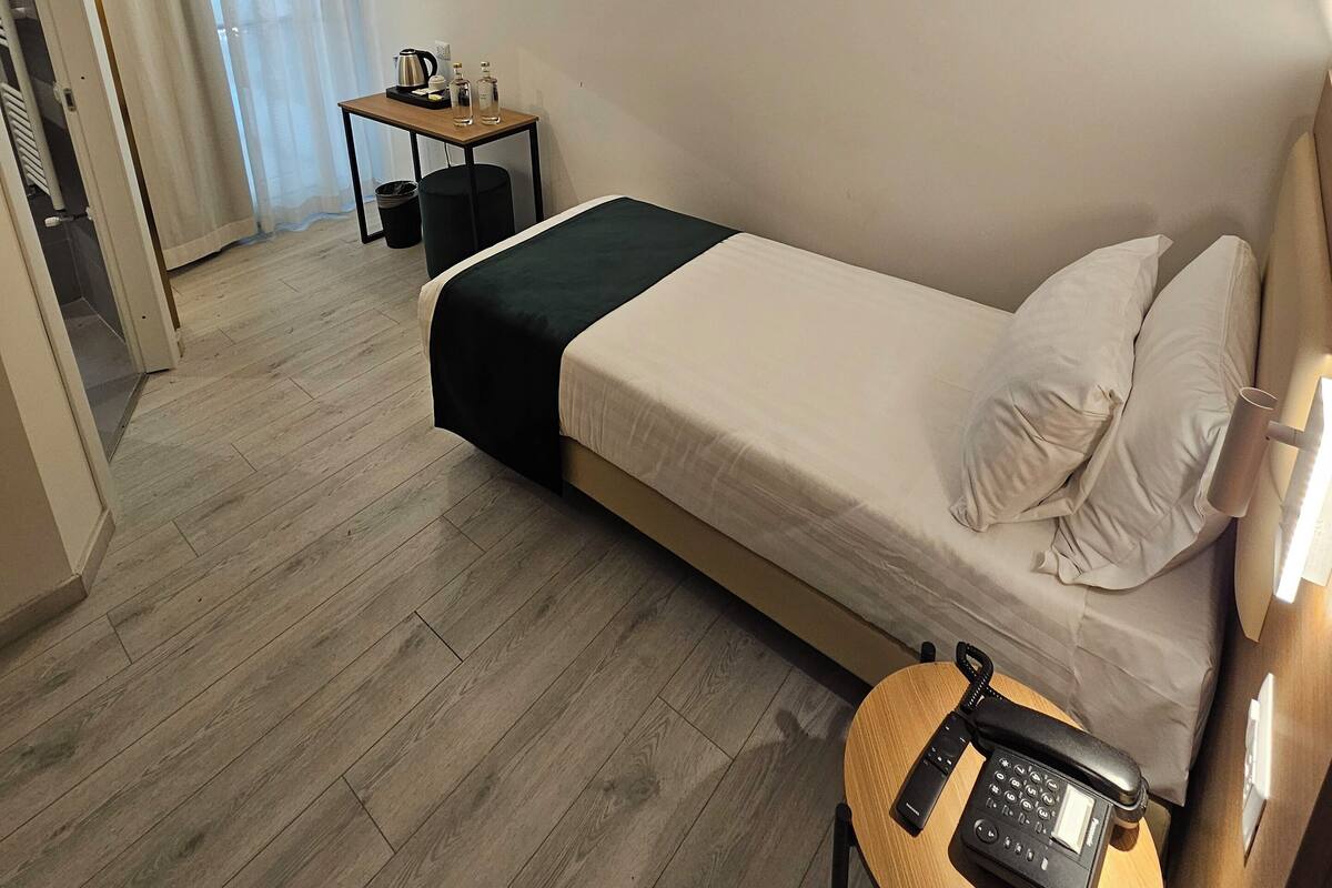 Ein Hotelzimmer mit Bett, Nachttisch mit Telefon, kleinem Tisch mit Wasserkocher und Tassen sowie einem Fenster mit Vorhängen.