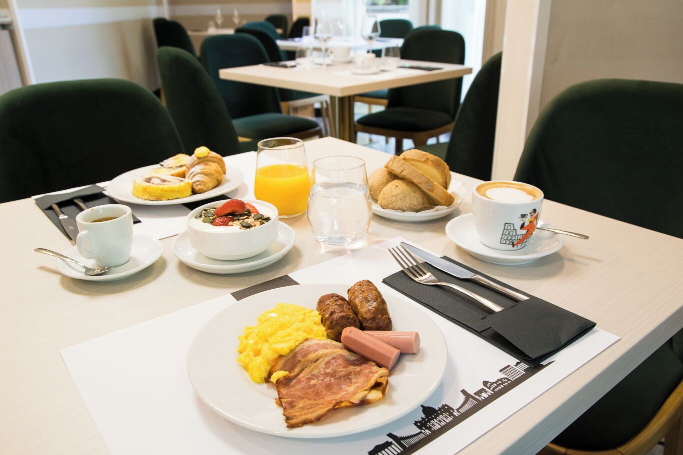 Un desayuno con huevos revueltos, salchichas, jamón, un tazón de cereal con fruta, un croissant, un vaso de jugo de naranja y una taza de café.