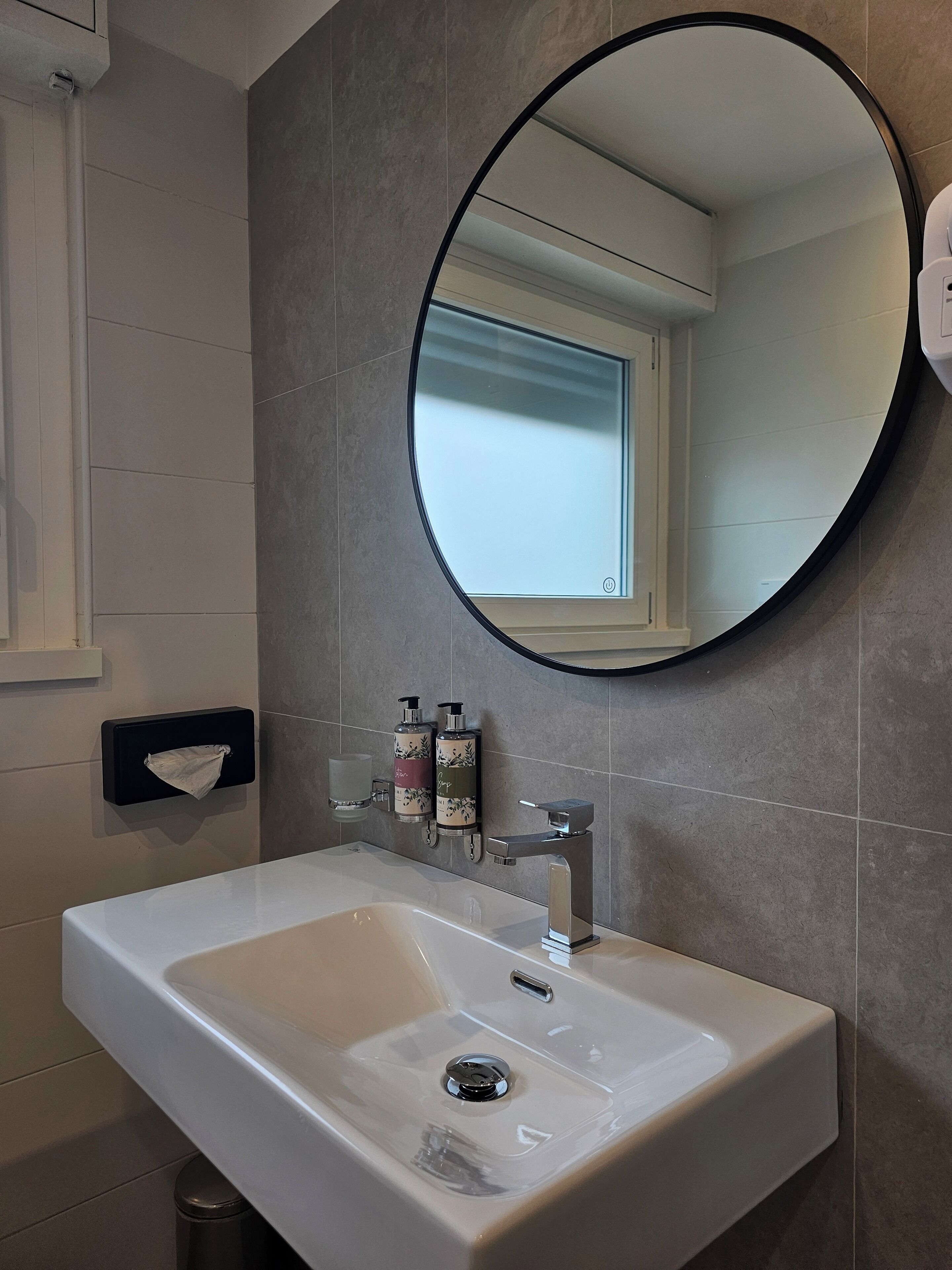 Un baño con un lavamanos blanco, un espejo redondo y dos botellas de jabón en la pared.