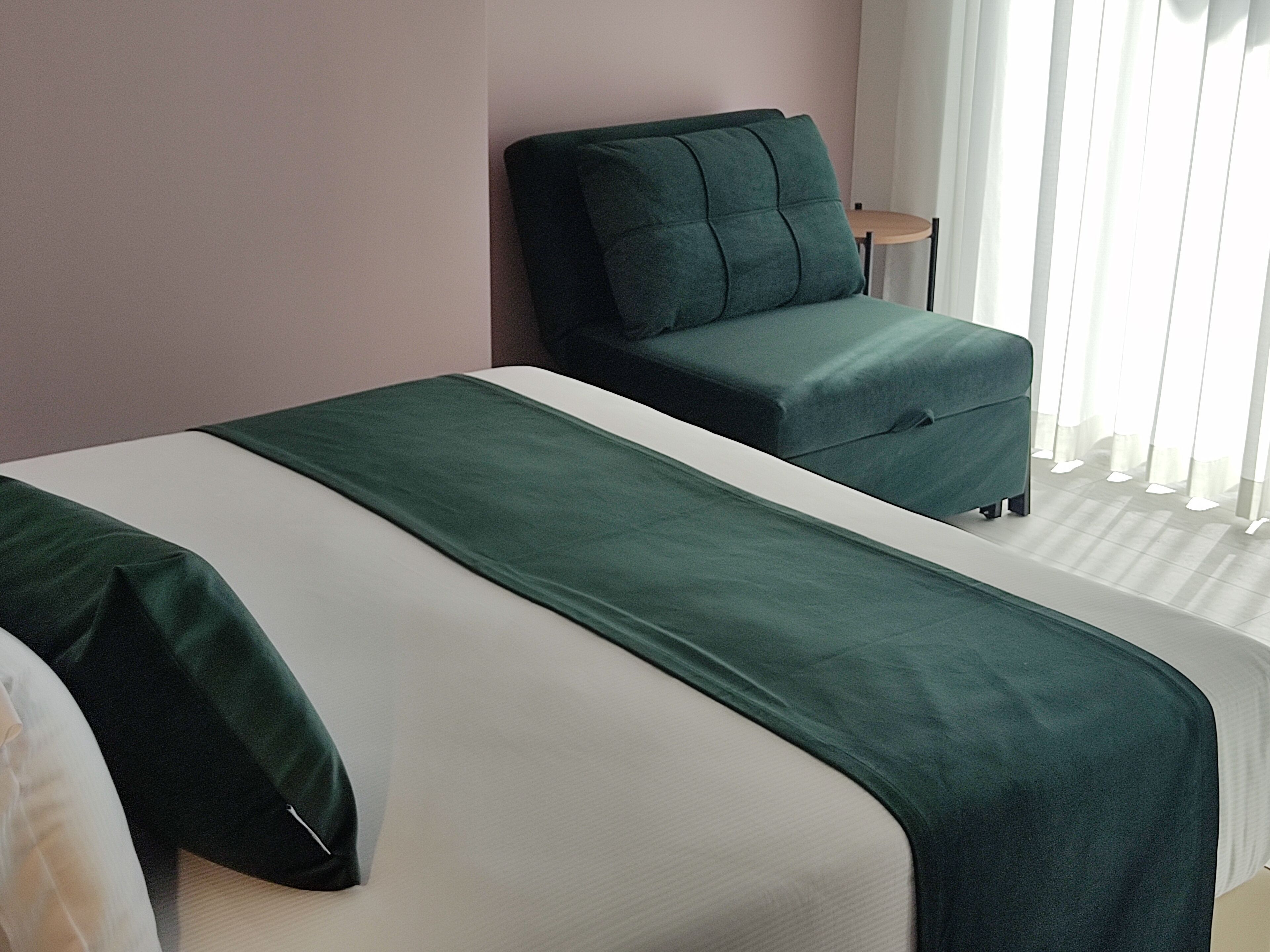 Habitación de hotel con dos camas, una silla tapizada de verde, una mesita y una ventana con cortinas translúcidas.