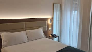 Habitación de hotel con una cama grande, una mesita de noche con teléfono, una lámpara de pared y una ventana con cortinas traslúcidas.