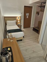 A hotel room with a bed, bedside table, and a wooden chair. There is a door with a sign on it, and a coat rack with hangers.