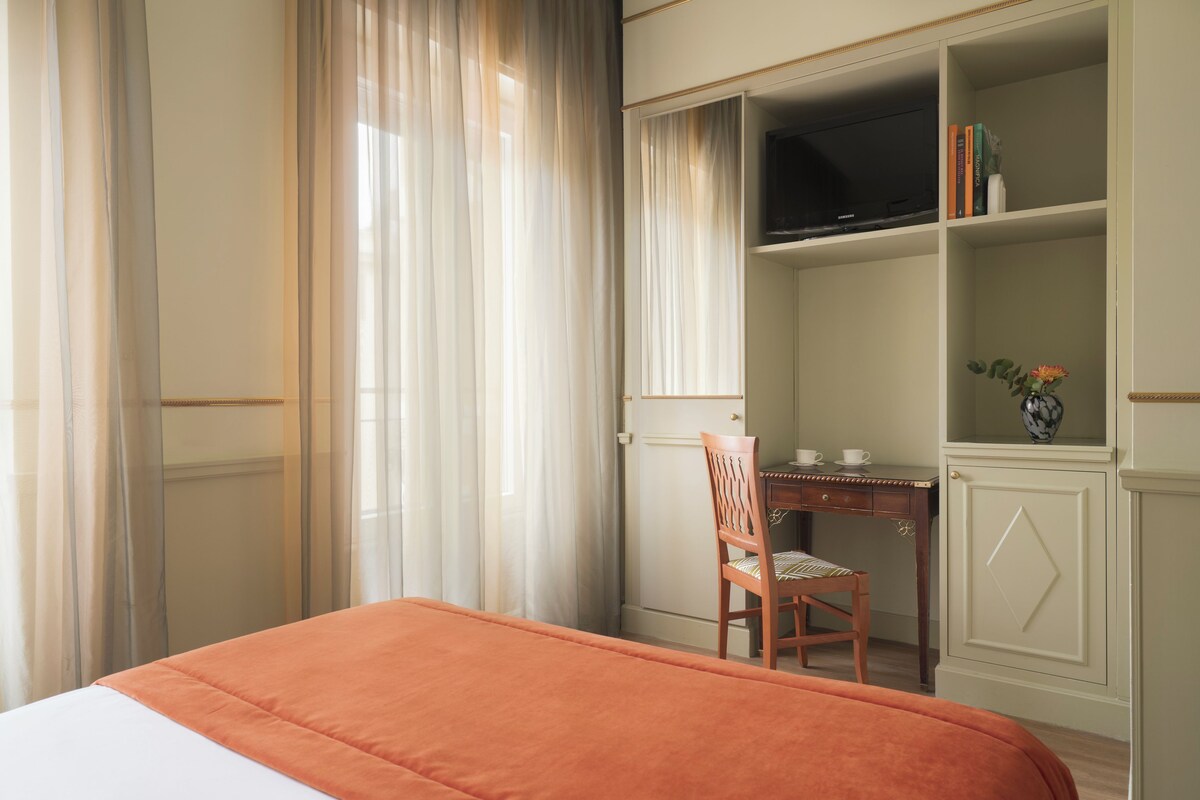 A bedroom with a bed, a desk with a chair, a television on a shelf, and a bookshelf.