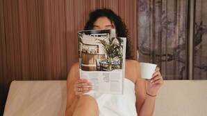 Uma pessoa sentada na cama, com uma revista, uma xícara e um prato de biscoitos.