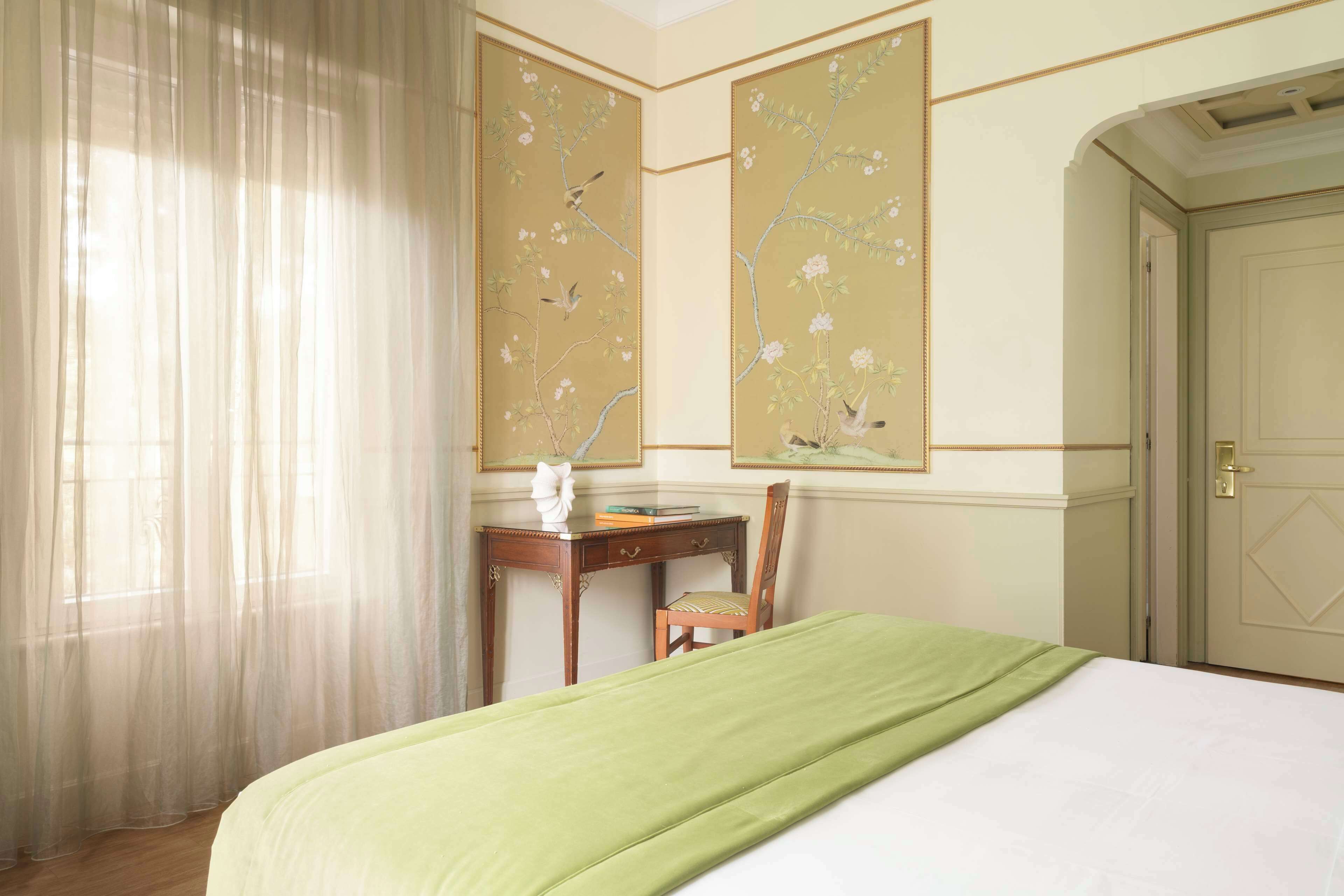 A hotel room with a bed, a desk, and a chair. There is a window with sheer curtains, and a door is visible in the background.