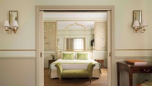 Quarto de hotel com uma cama grande, um banquinho, uma mesinha de cabeceira, um espelho e arandelas na parede.