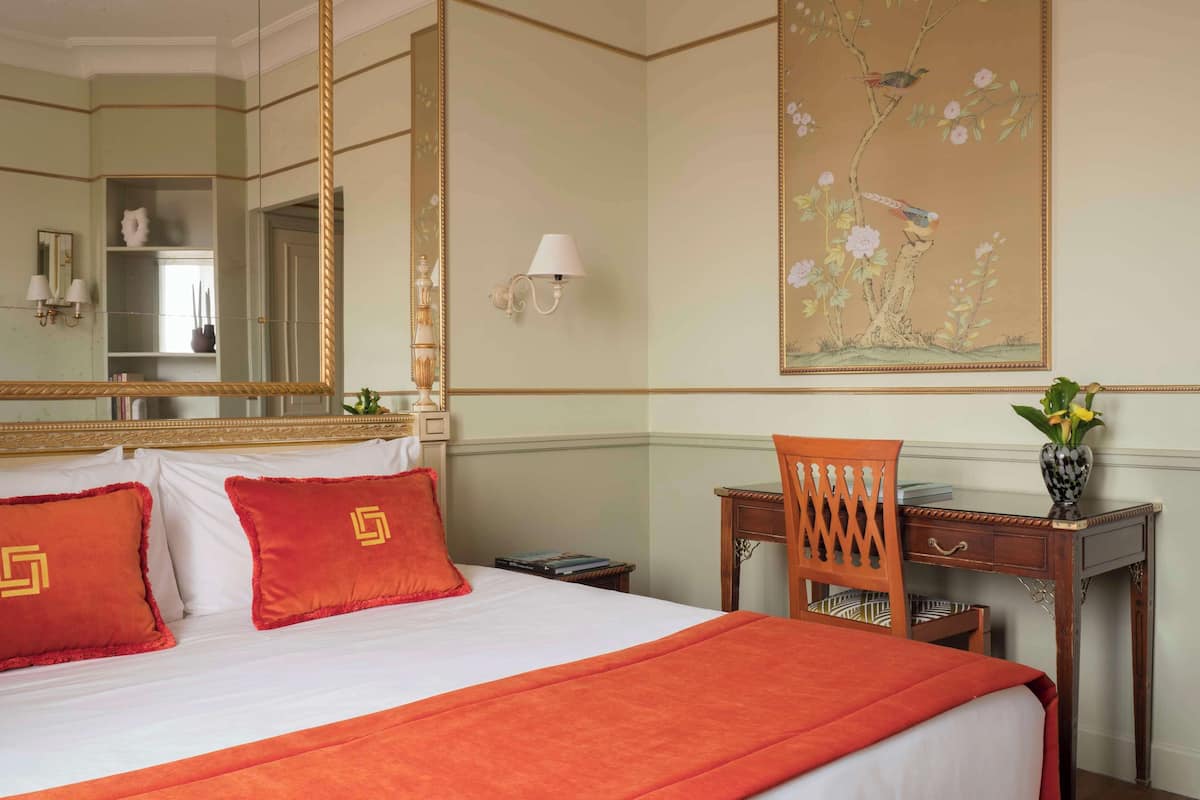 A hotel room with a large bed, a desk with a chair, a mirror, and a wall painting.