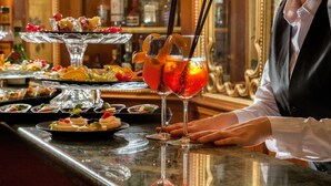 Uma área de bar com um bartender, um copo de drink vermelho com guarnição, e diversos doces em exposição.