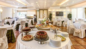 A banquet hall with round tables covered with white tablecloths, decorated with various desserts and fruit platters. - Appia Park Hotel (Rome)
