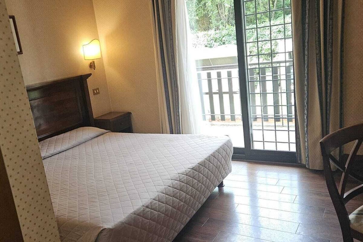 A bedroom with a bed, a nightstand, a chair, a window with a view of greenery, and a wall-mounted light.