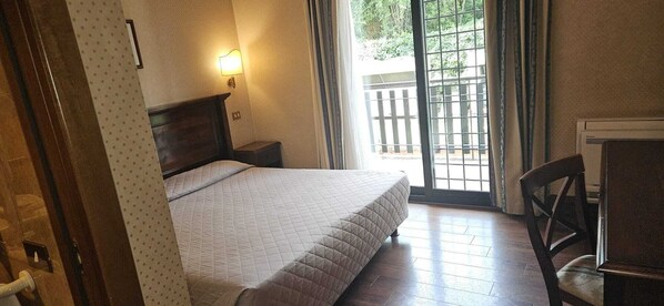 A bedroom with a bed, a nightstand, a chair, a window with a view of greenery, and a wall-mounted light. - Appia Park Hotel (Rome)